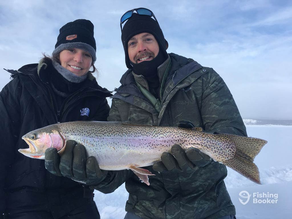 Two people dressed in winter clothing are holding a large Trout on a snowy landscape, showing off the excitement of Colorado's fishing seasons. Both anglers can't hide their smiles as they proudly display their catch for the camera.