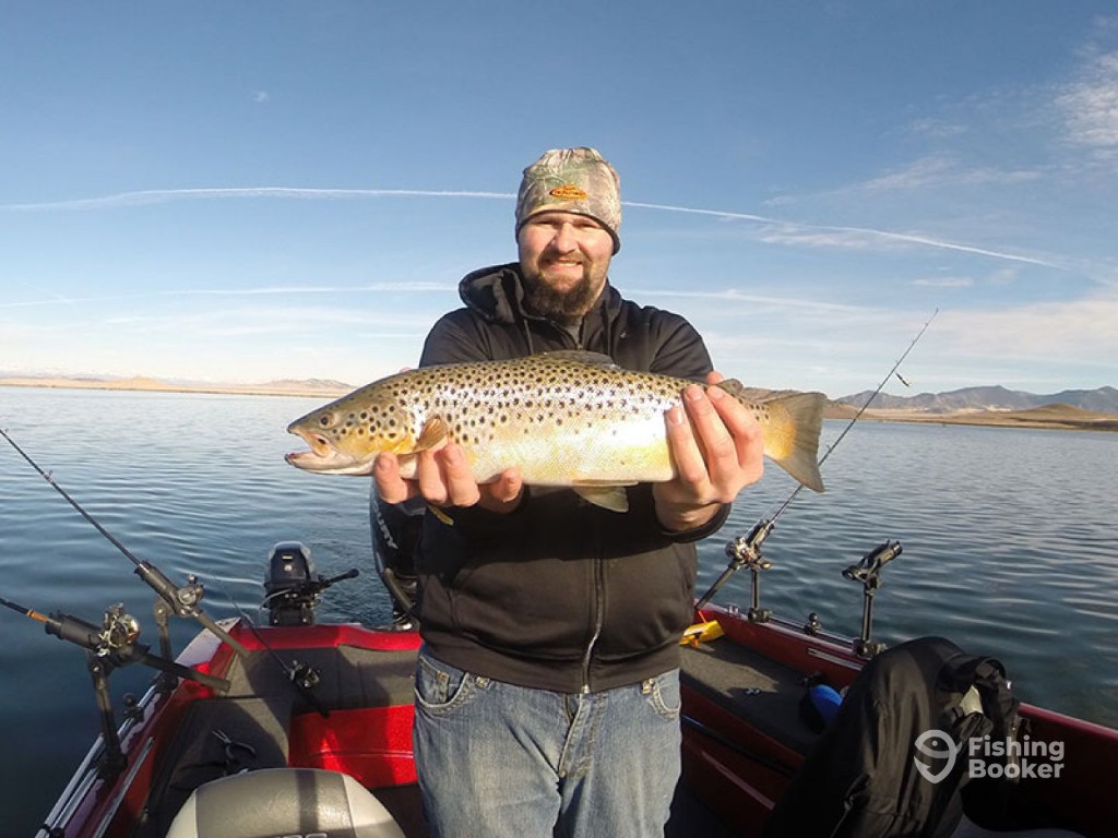 A man standing on a boat holds a large Brown Trout with both hands, with calm waters visible behind him on a clear day