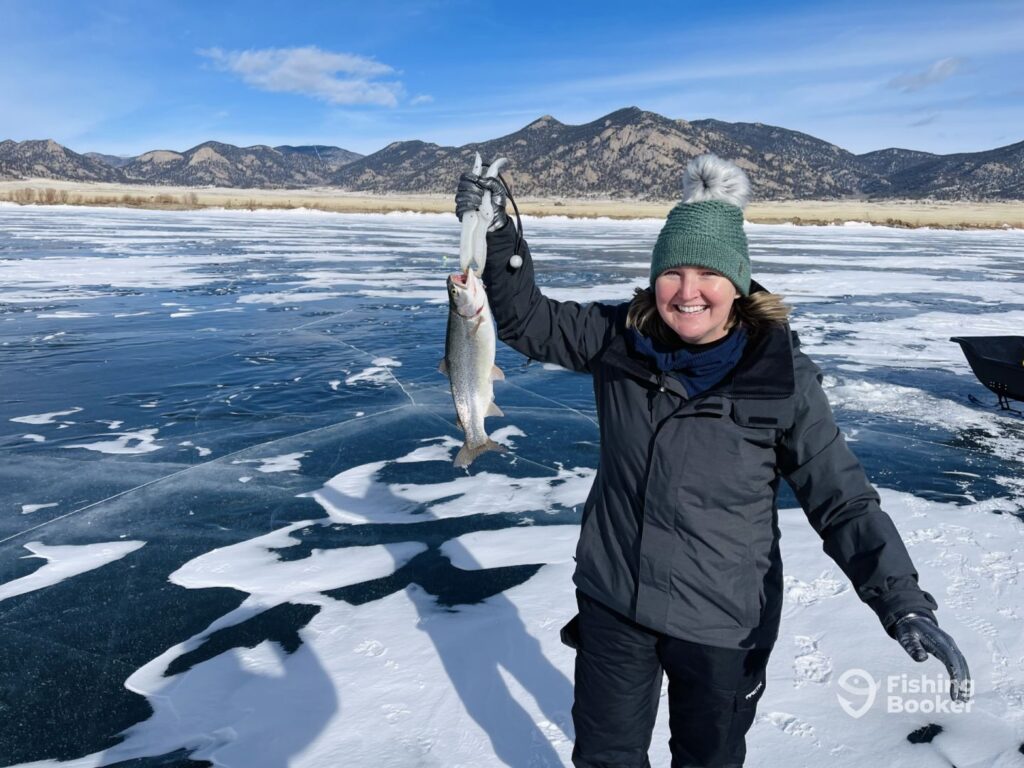 A woman dressed in winter clothing stands on a frozen lake during Colorado's fishing season, holding up a caught Trout with majestic mountains and a bright blue sky in the background.