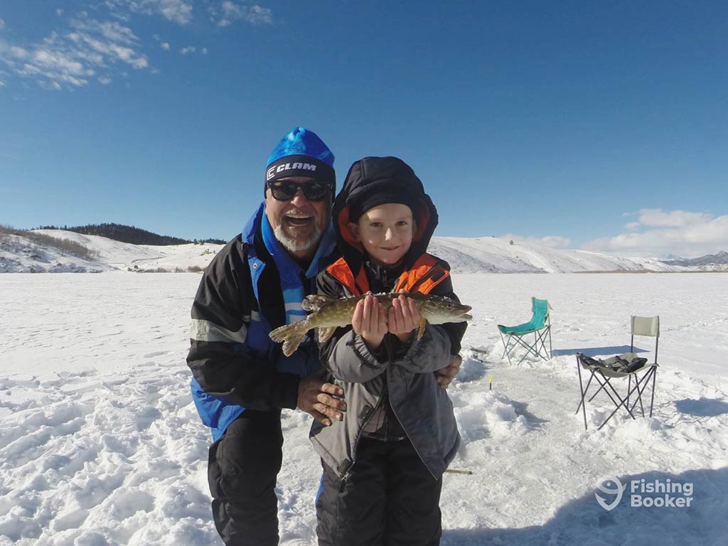 An adult and a child pose on a snowy landscape in Colorado, with the child proudly holding a Trout. Portable chairs and fishing gear are visible in the background, suggesting they've been enjoying several hours of cold-weather angling together.