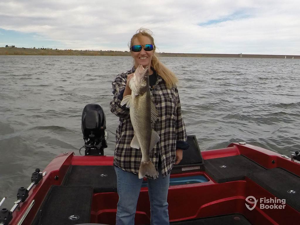A woman on a red boat holds up a large Walleye, smiling, with a scenic lake and cloudy sky behind her.