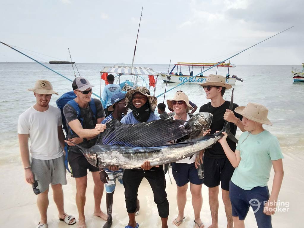 A group of anglers on a beach in Kenya, smiling as they hold a large Sailfish with a fishing boat and the ocean visible behind them on a cloudy day.