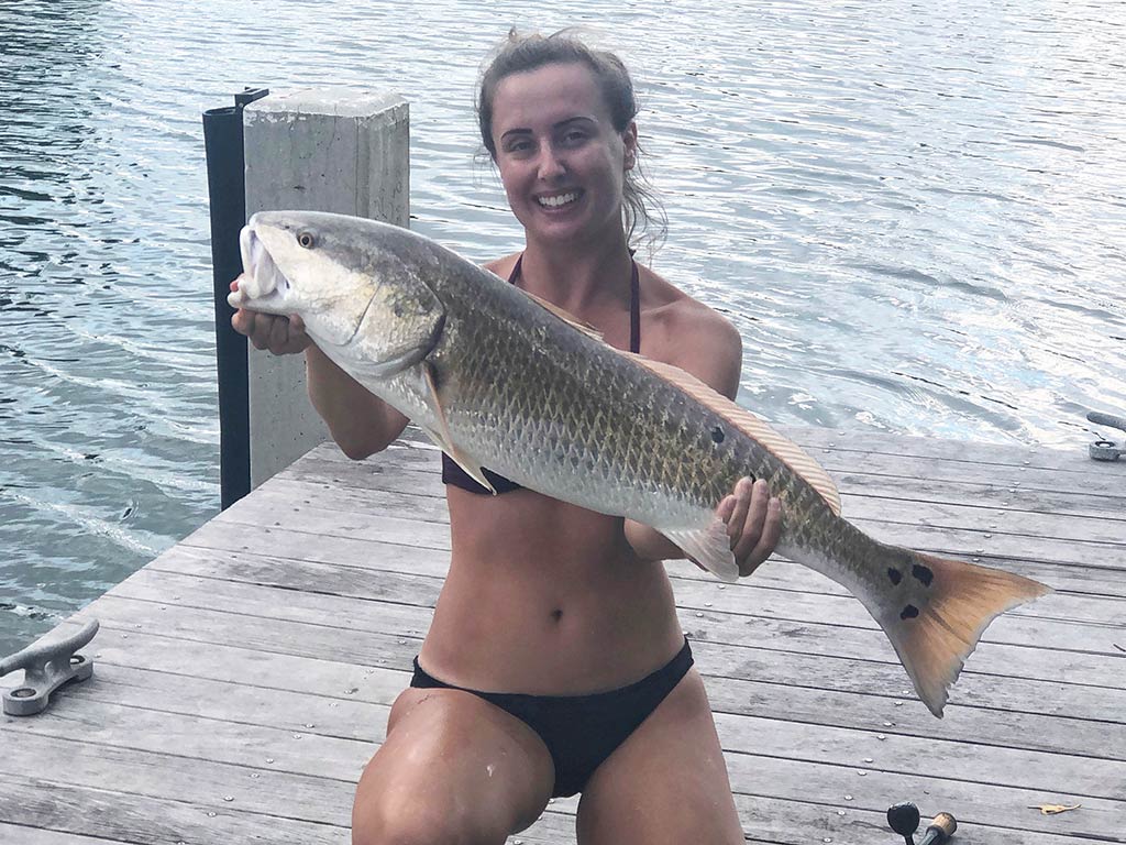 A woman kneeling on a dock with a fishing rod by her side as she holds a big Redfish on a gray day.