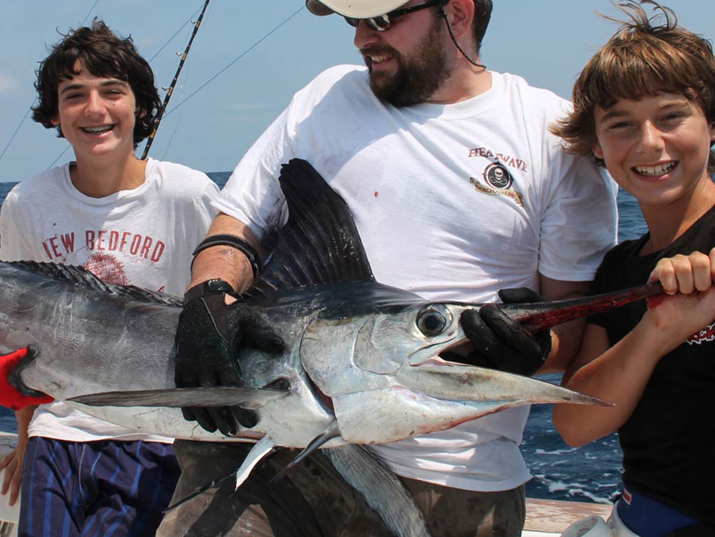 A man sits between two children on a fishing charter with a White Marlin across their laps.