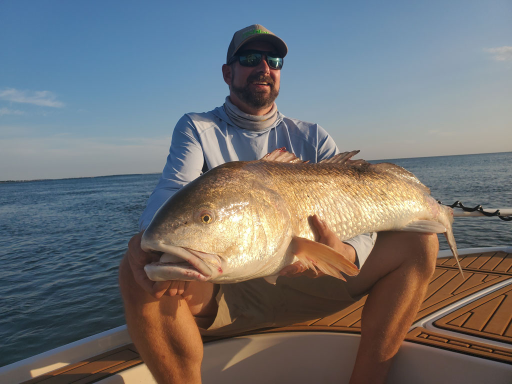A man on a fishing charter with a large Bull Redfish across his lap on the open waters of the Atlantic Ocean on a clear day.