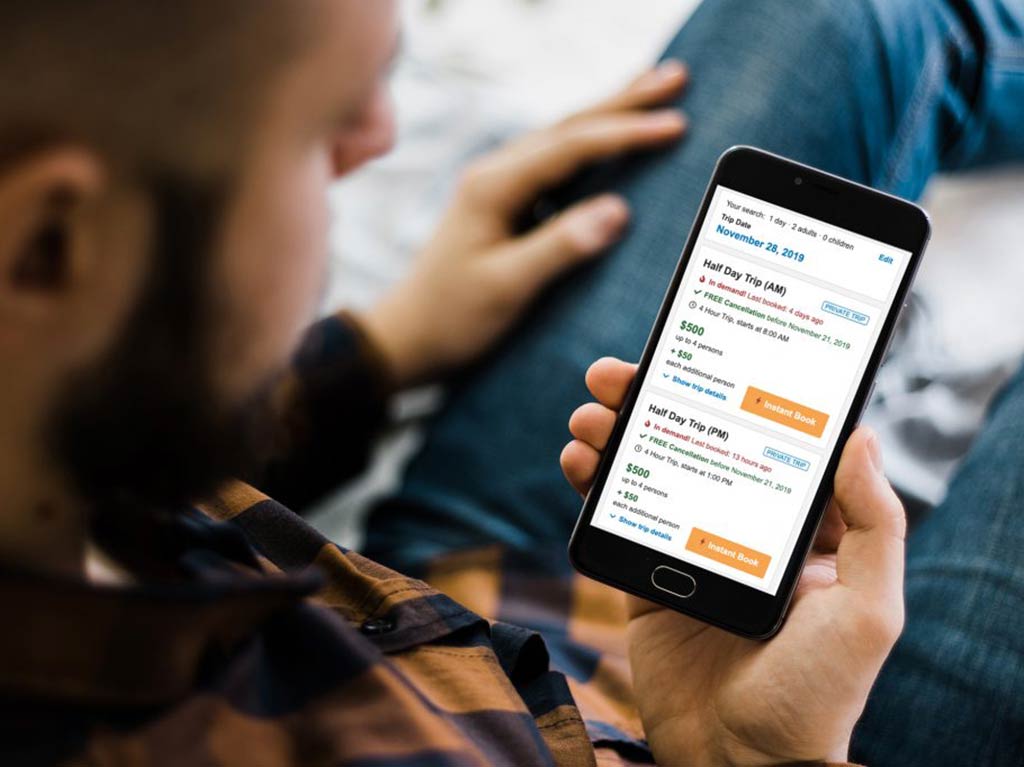 Вид сверху на мобильный телефон, показывающий варианты поездок в приложении FishingBooker, на фоне размытых фигур человека.
