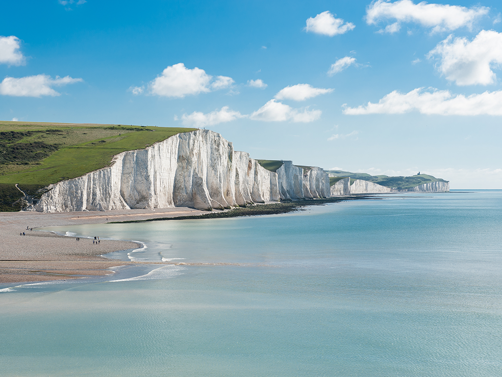A view of the White Cliffs of Dover in the UK, with a beach visible on the left of the image and people walking on it on a clear day.