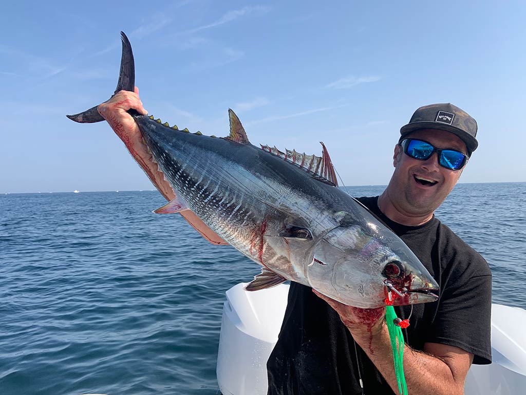 A man in a baseball cap and sunglasses standing on a boat on a clear day, holfing a large Tuna with a lure hanging out of its mouth. 