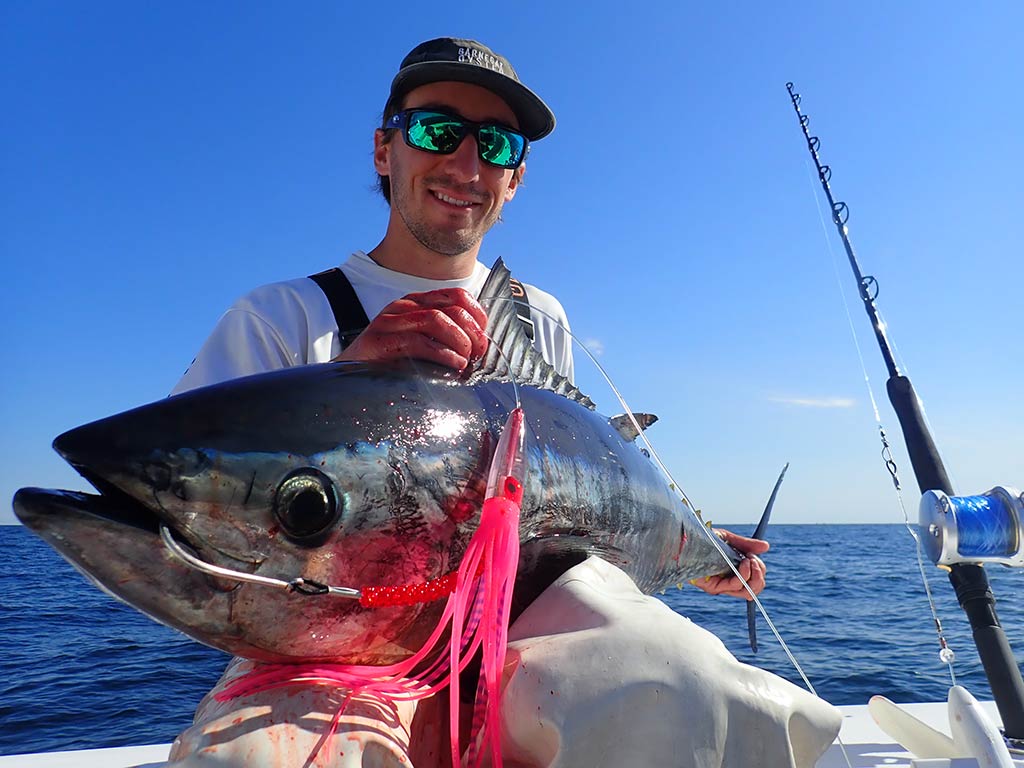 An angler in sunglasses and a baseball cap sits on the side of a fishing boat with a Tuna across their lap, with a lure hanging out of the fish's mouth.