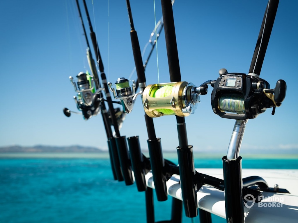 Four fishing rods with reels are mounted on a boat, overlooking clear blue ocean water with a distant shoreline visible under a clear sky.