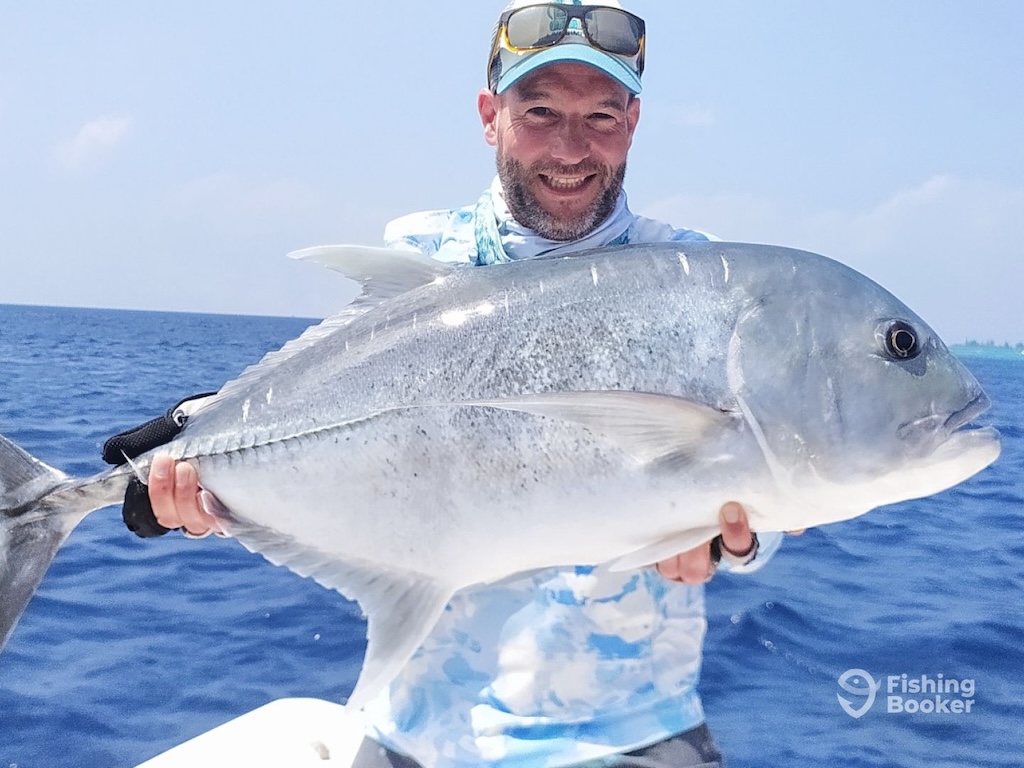 A man wearing a hat and sunglasses while holding a large Giant Trevally on a boat, with the sea and sky behind them.