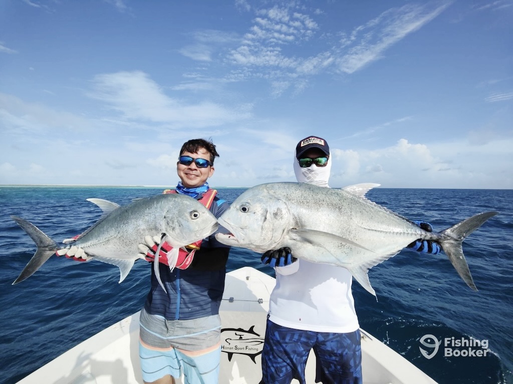 Two people on a boat holding large Giant Trevallies, with the ocean and blue sky in the background. 