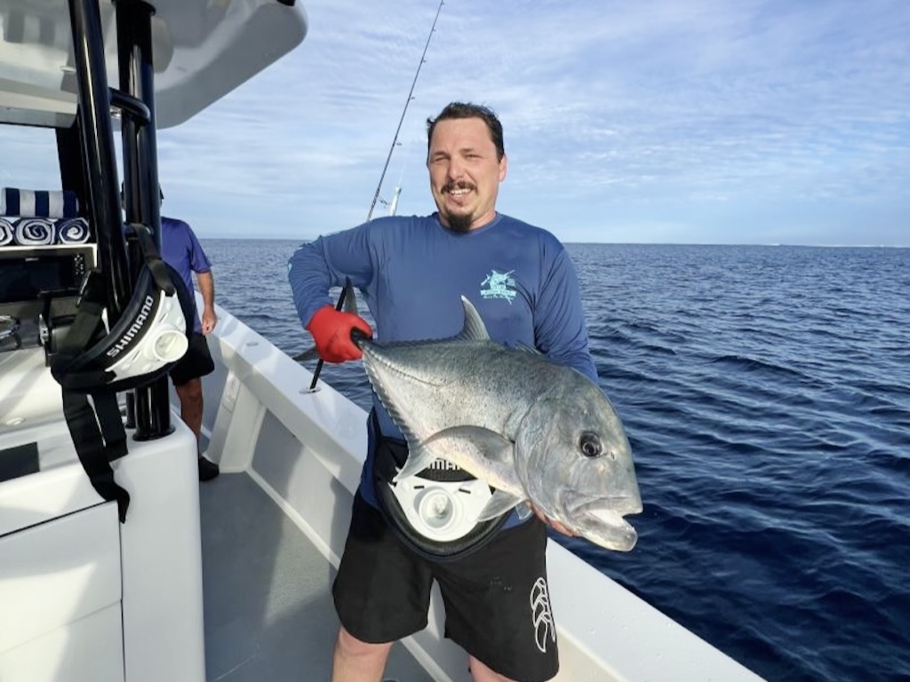 A man on a boat holding a big GT, with the ocean and a fishing rod in the background.