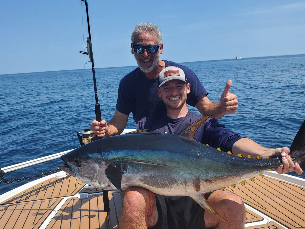 Two men on a boat pose with a large Tuna; one’s holding the fish while the other is giving a thumbs up. The ocean and blue sky are in the background.