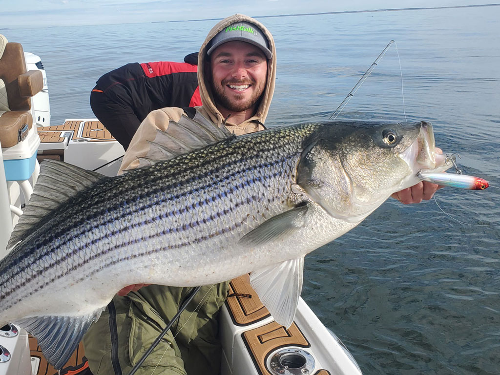 Мужчина в лодке держит крупного окуня с приманкой в пасти, улыбается на камеру. На заднем плане вода