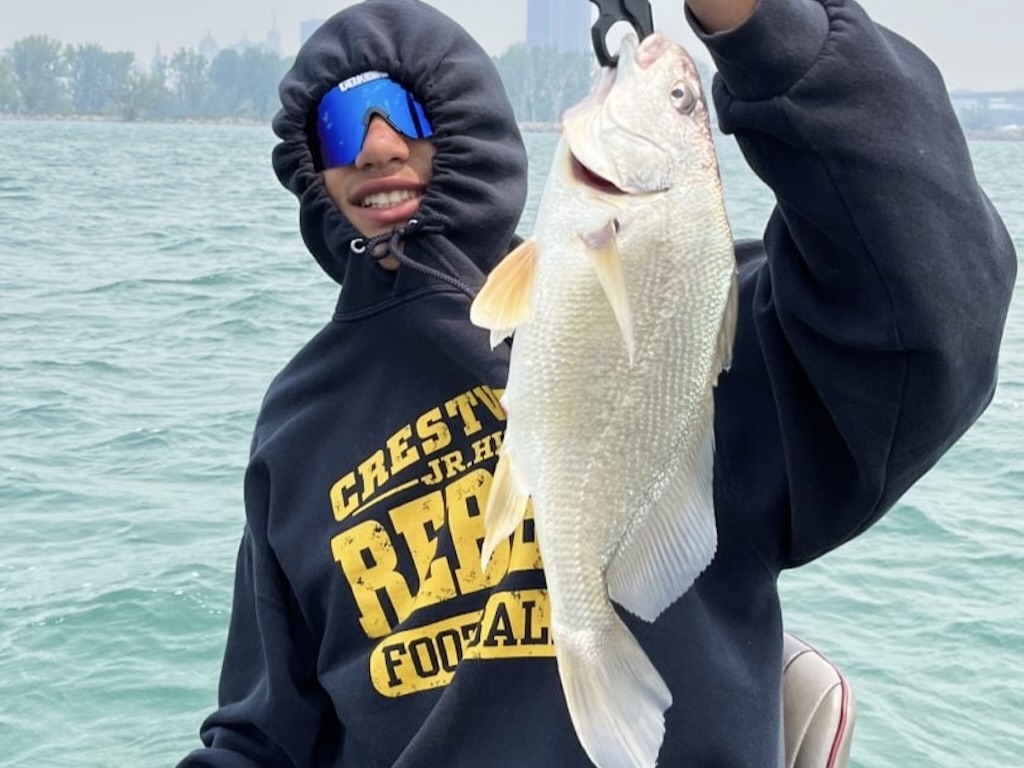 Someone wearing sunglasses and a hoodie as they hold up a large Freshwater Drum above the water on a boat. You can see trees and a city skyline in the background.