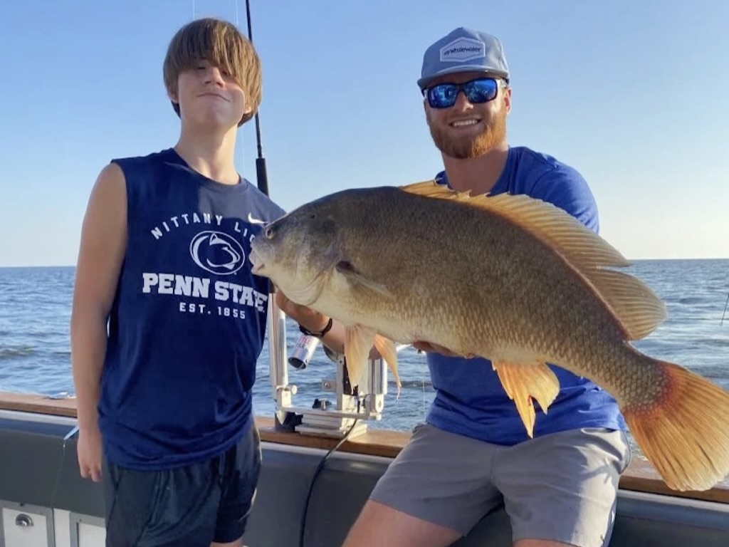 Two people on a boat are holding a big Freshwater Drum together, with water and sky behind them. One is wearing a Penn State sleeveless shirt, while the other is wearing got sunglasses and a cap.
