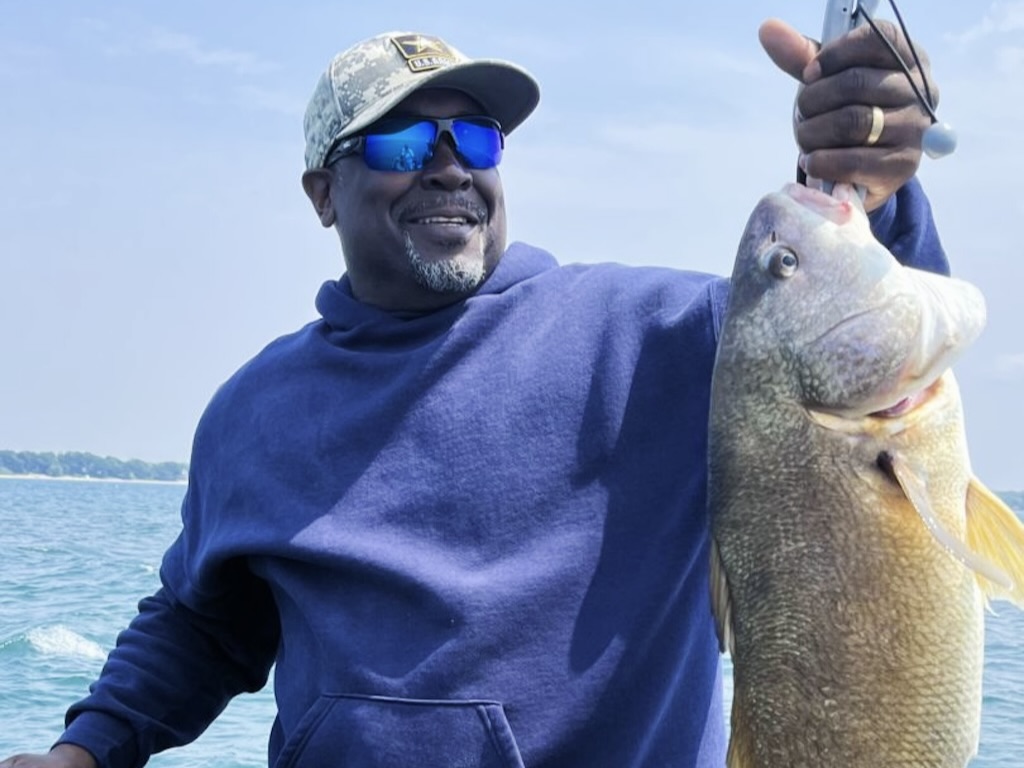 A man wearing a blue hoodie and cap as he holds up a large Freshwater Drum while standing on a boat in open water.