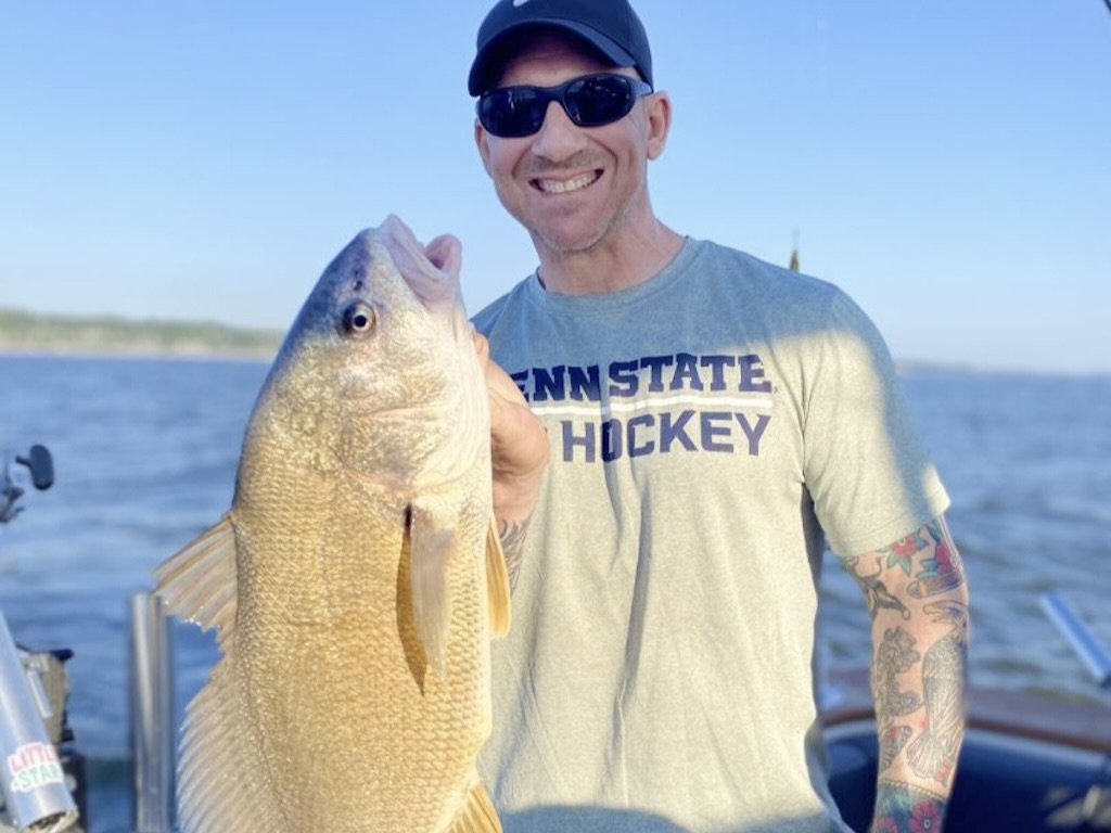 A man wearing sunglasses and a Penn State Hockey shirt proudly holds up a large Freshwater Drum on a boat, with water and blue sky in the background.