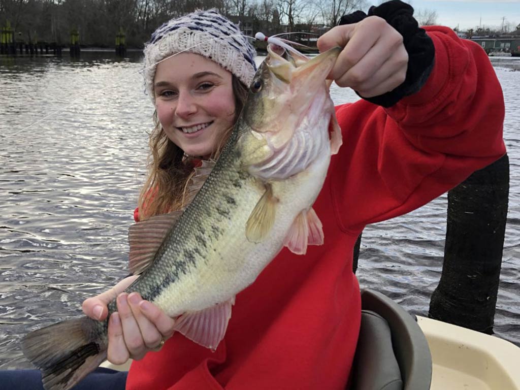 A girl in a red jacket and knit hat, holding a large Bass, with water and trees behind her.