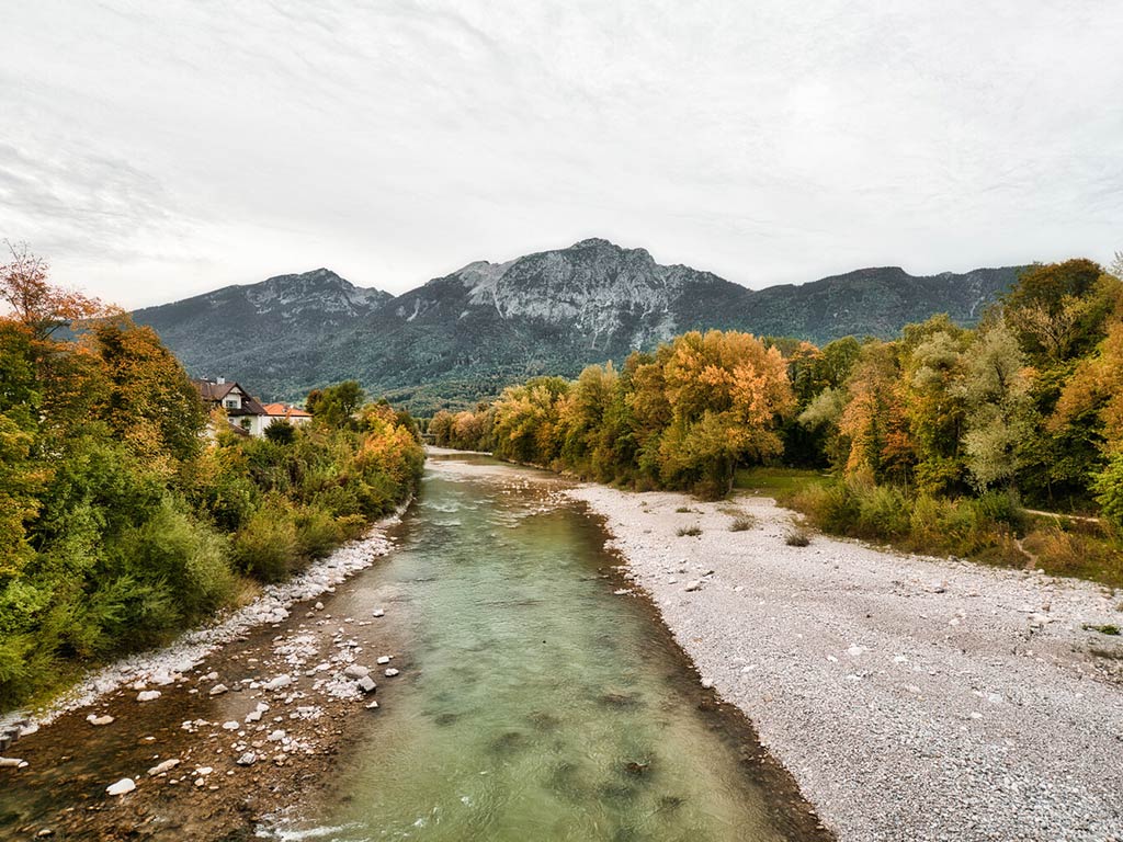 A shallow river with rocky banks flows through a valley lined with autumn trees, while mountains and a cloudy sky are in the background. 