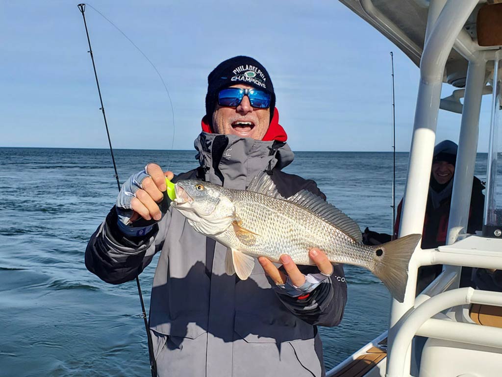 A man wearing sunglasses and a hat, holding a large Redfish on a boat, with the ocean in the background.