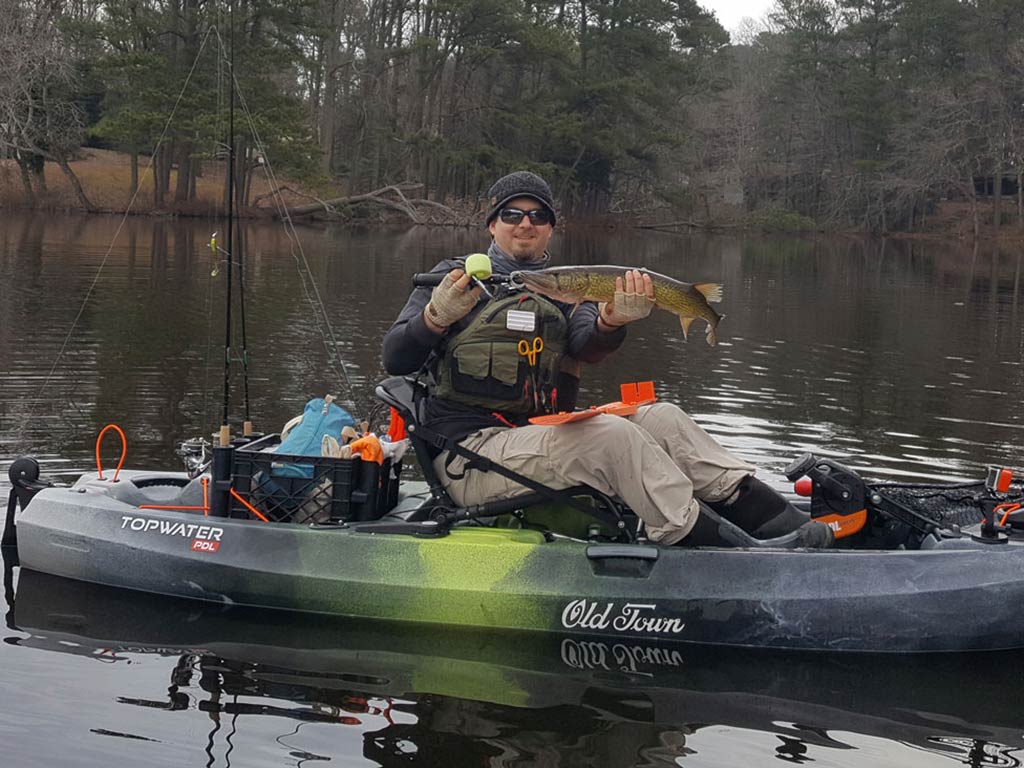 A man sitting in a kayak on a lake, holding up a Pickerel they’ve caught, with fishing gear scattered around and trees in the background.