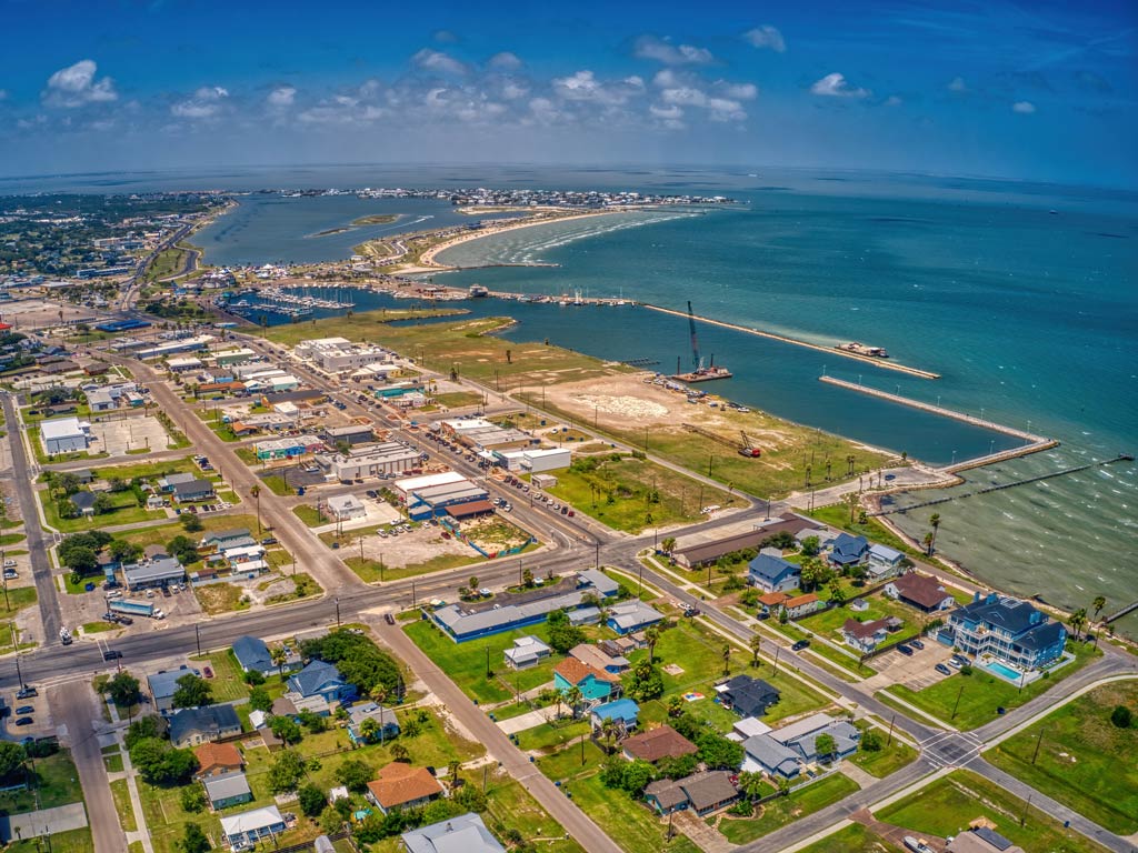 An aerial view of a coastal town in Texas called Rockport, with homes, roads, a marina, and docks stretching into blue-green water. 