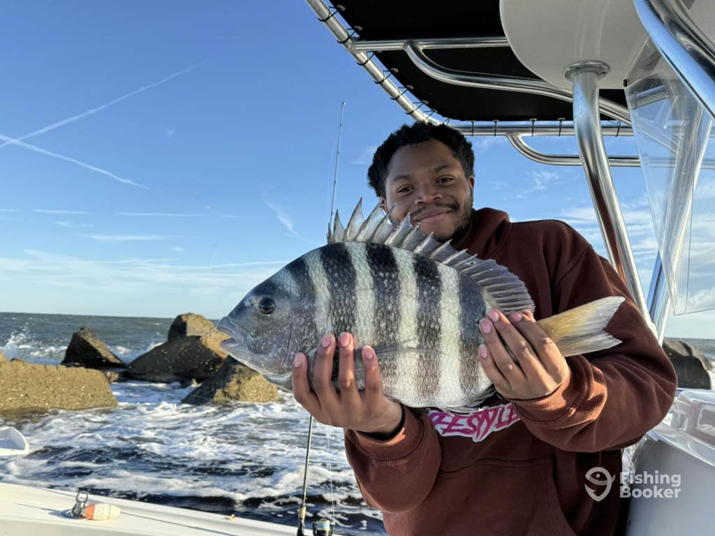 An angler standing on a charter boat in Jacksonville and posing with a large Sheepshead, with rocks and ocean waves behind them.