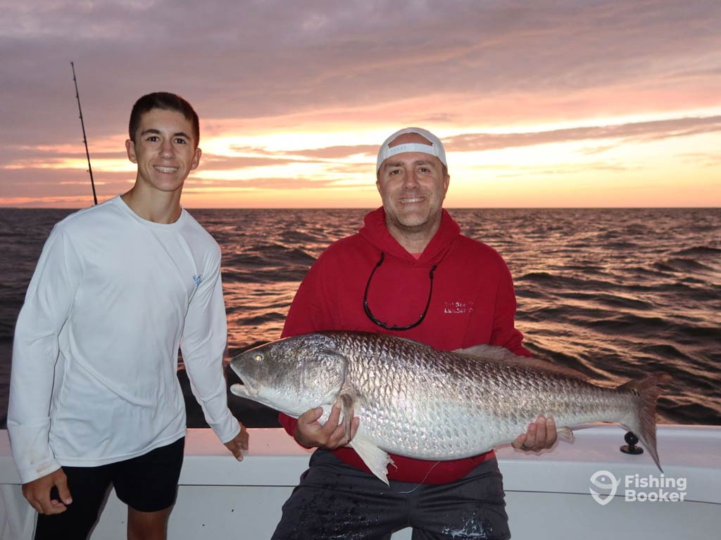 Two anglers are on a boat at sunset. The person on the right is holding a large Redfish, while the person on the left stands and smiles. The ocean and sky are in the background while they pose in Morehead City, one of the winter fishing destinations every angler should visit.
