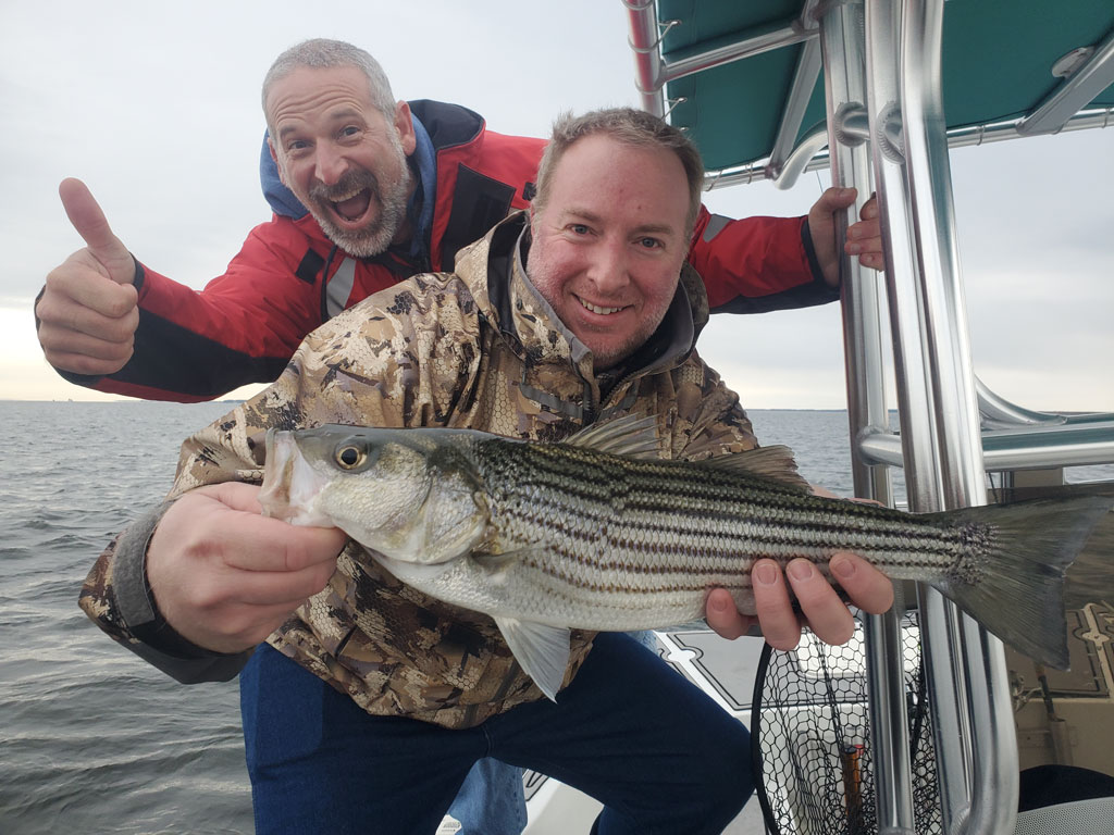 Two men are on a boat –one is holding a Striped Bass fish while the other stands behind him giving a thumbs-up. They're both clearly enjoying their fishing trip.