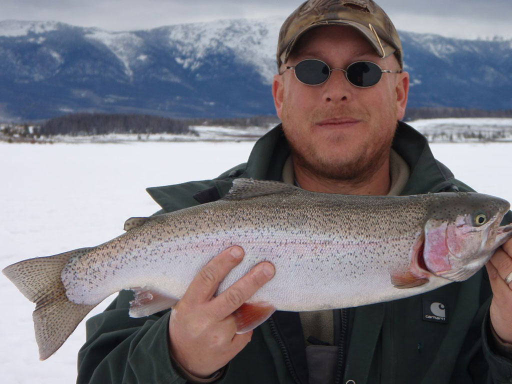 A man wearing sunglasses and a cap holds a large Trout in front of a snowy landscape with mountains in the background.
