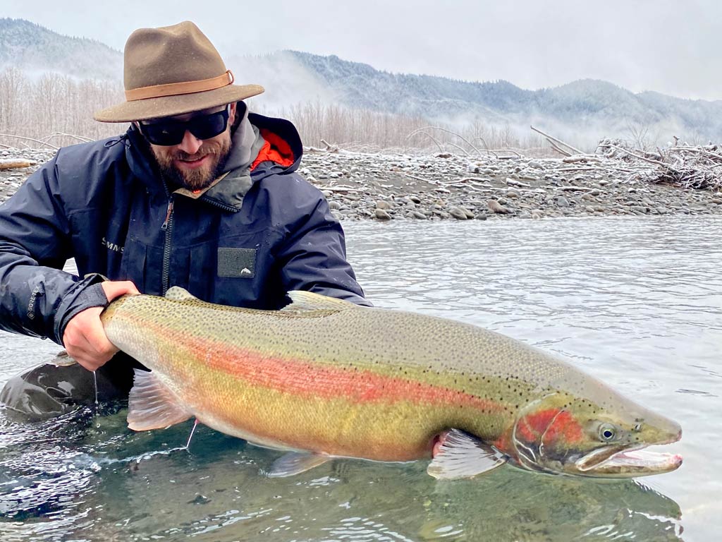 Someone in a hat and sunglasses holds a large Rainbow Trout in shallow water, showing off one of the top winter fishing spots with snowy trees and mountains behind.