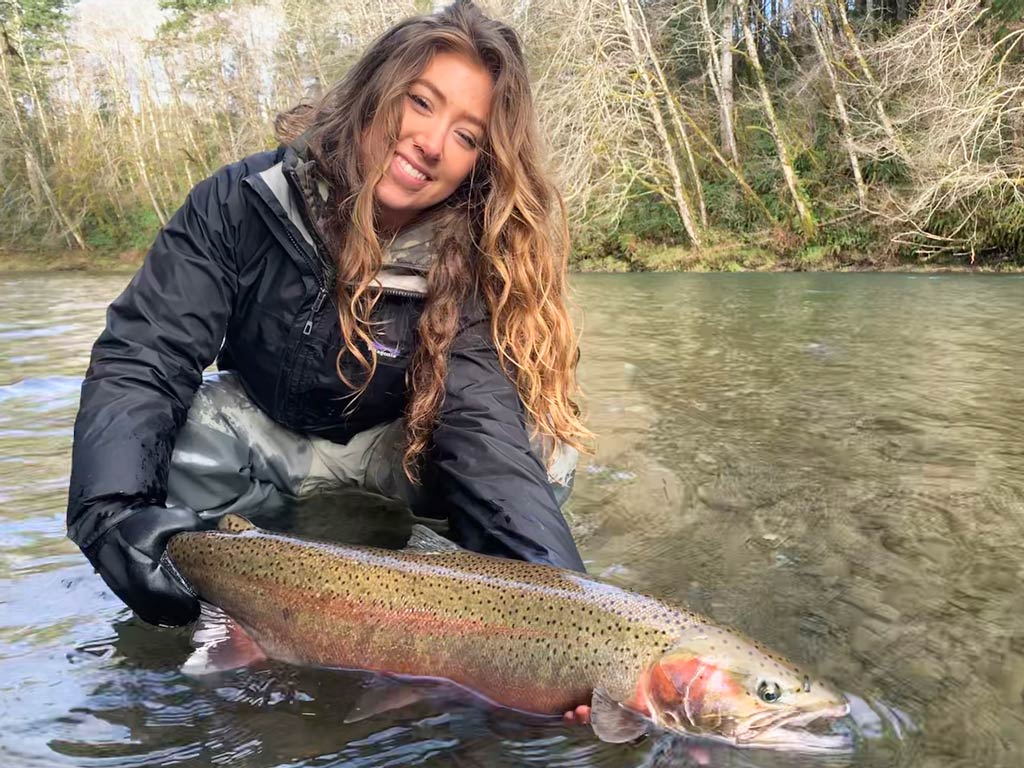A female angler in waders, kneeling in a shallow river and holding a large fish - it's the perfect spot for a winter fishing destination. Trees and greenery can be seen in the background.