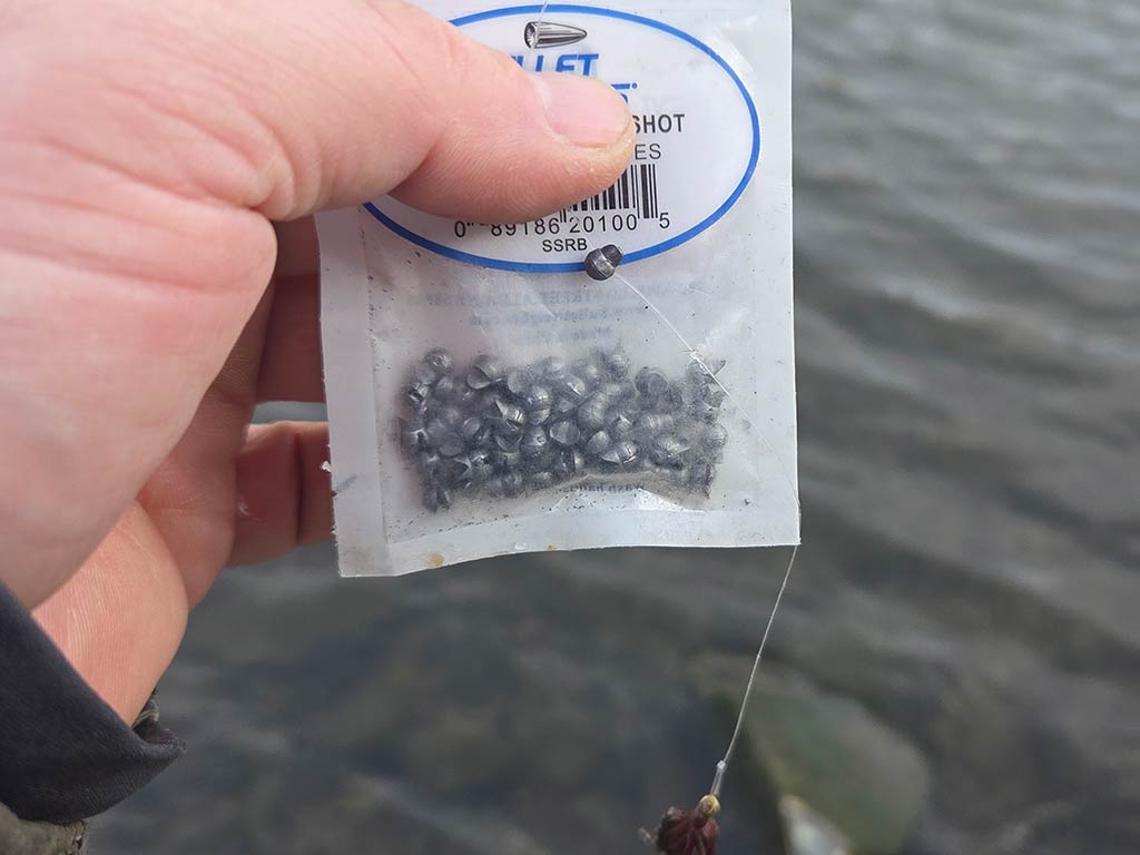A hand holding a small, clear plastic bag with miniature fishing sinkers near a body of water.