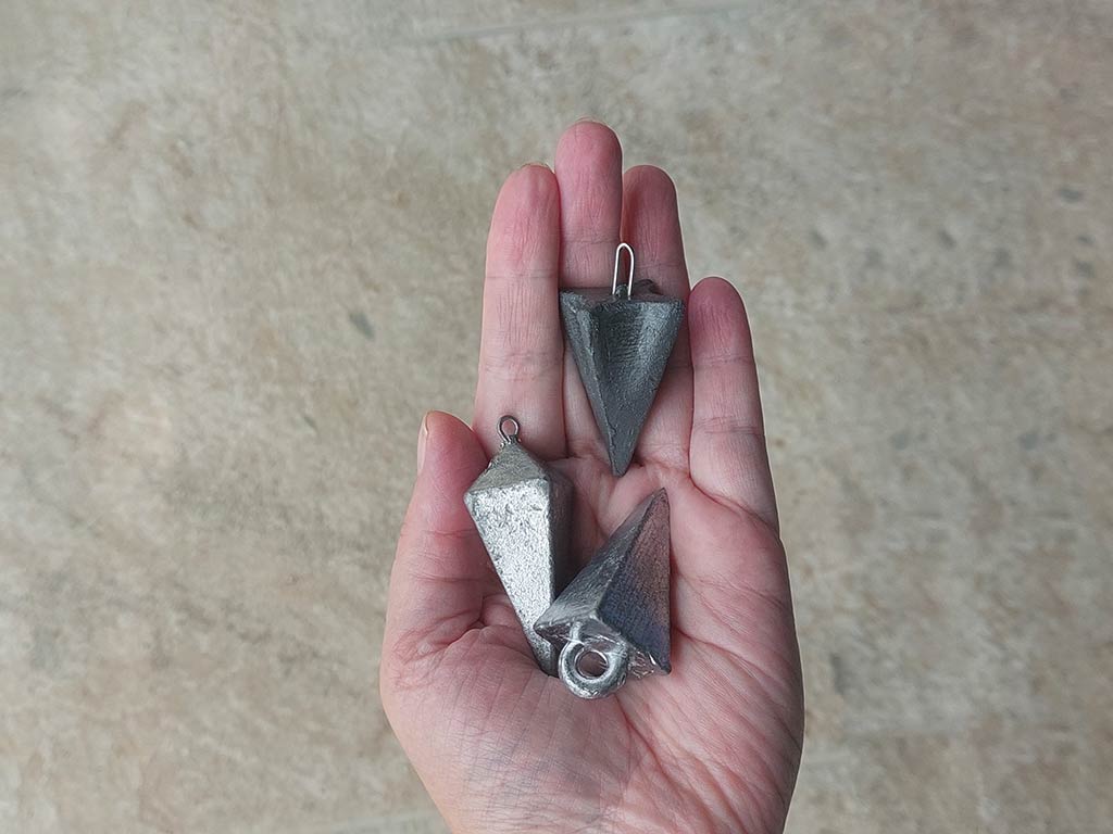 A hand holding three gray, metal pyramid fishing sinkers against a sandy background.