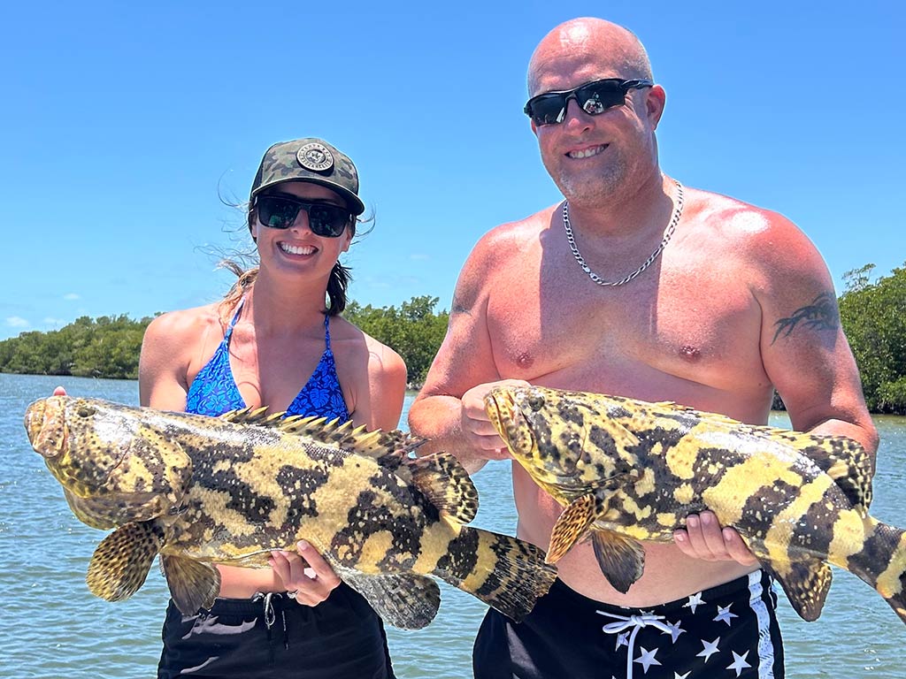 Two people standing outdoors, each holding a large, patterned Gag Grouper with water and greenery in the background under a clear blue sky.