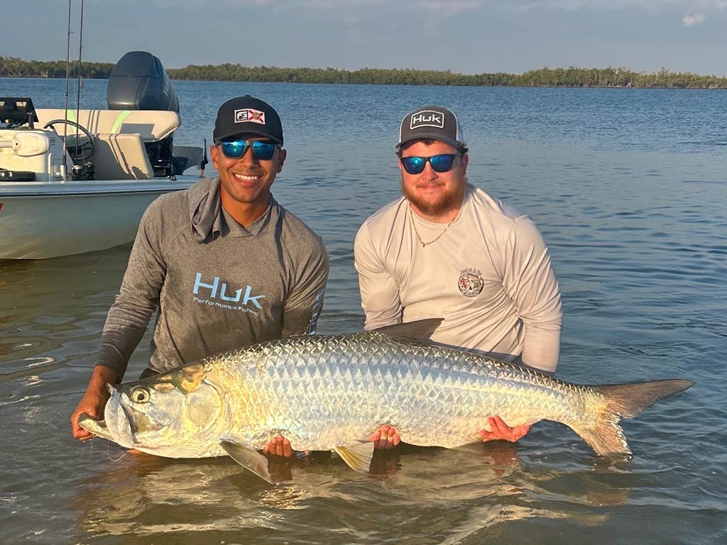 Two men standing in shallow water are holding a large, silver Tarpon with a boat and shoreline visible in the background.