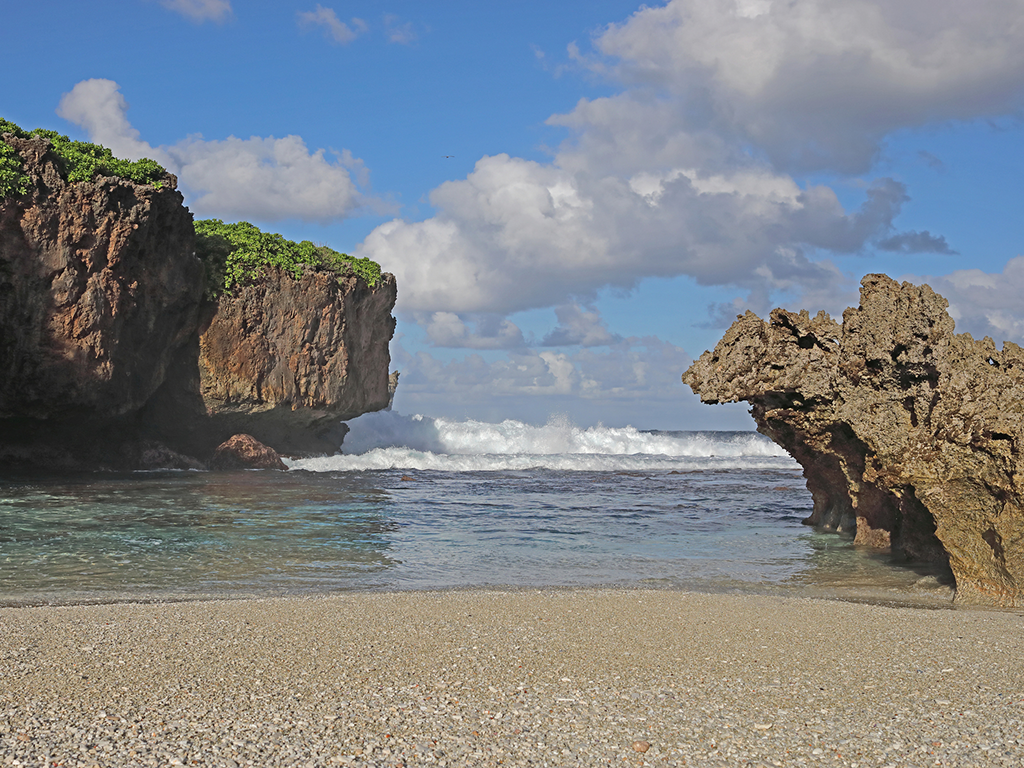 Rocky cliffs and clear waves border a pebbled beach under a blue sky with scattered clouds.
