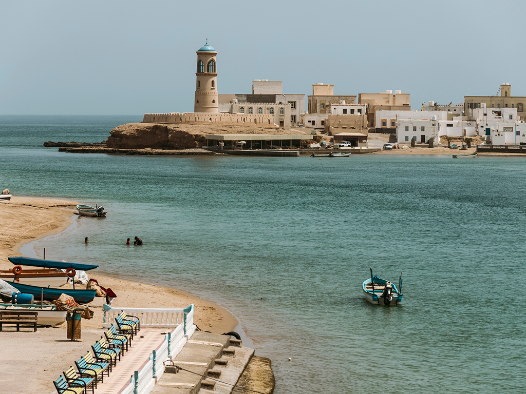 A coastal town in Oman with white buildings, an oriental lighthouse, and boats on calm blue water and a sandy beach with empty benches in the foreground.