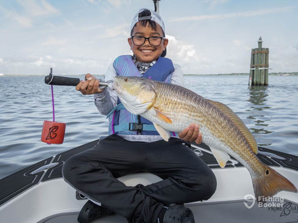 A photo featuring a happy kid sitting on a charter boat and posing with Redfish caught in Houston on a cloudy day.