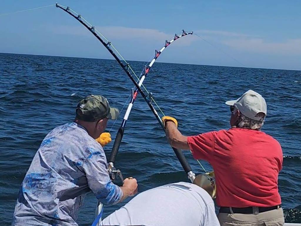 A view from behind of two men with crossed rods as another man bends down behind them, battling a Tuna each in the deep waters of the Atlantic on a clear day.