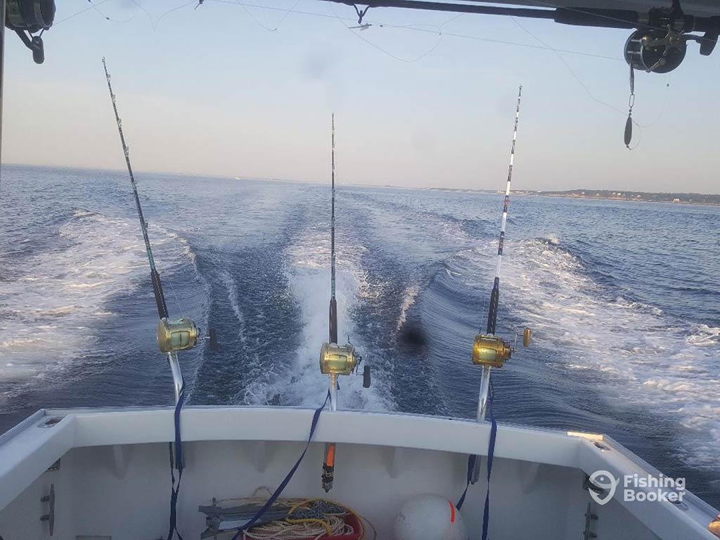 A view out of the back of a deep sea fishing boat, with three trolling rods set up to trail behind a boat with the boat's wake visible in the water near sunset on a clear day.