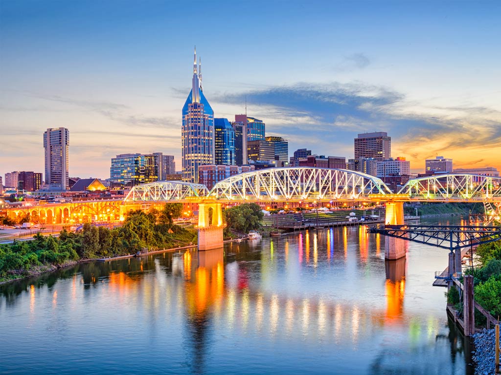 A photo featuring the city of Nashville dotted with tall buildings as seen from the river during the evening.