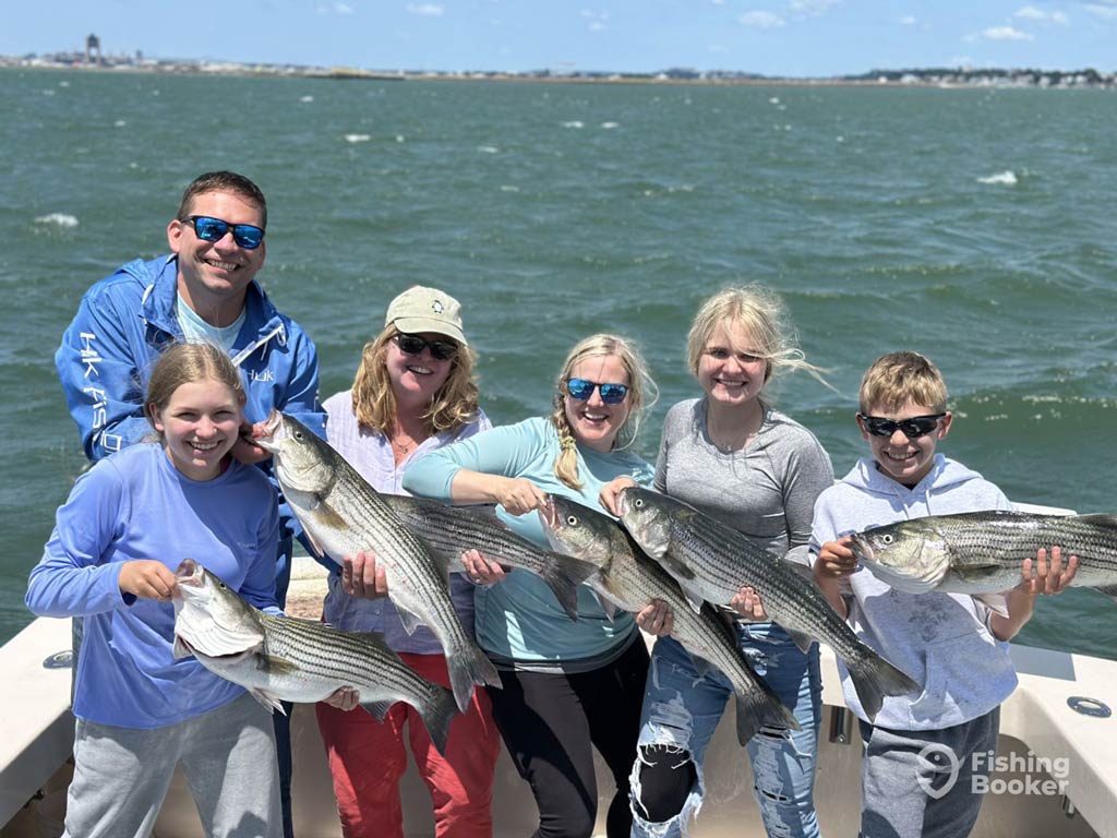 A photo featuring a family happily posing on a charter boat on a sunny day with a lot of Striped Bass catches they reeled in on their angling trip in Boston.