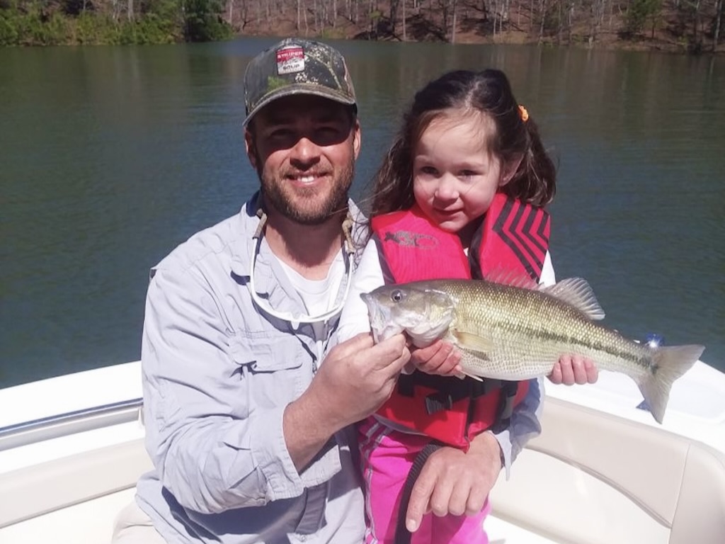 A father and daughter crouching on a boat while holding a Spotted Bass that they caught from a calm lake.
