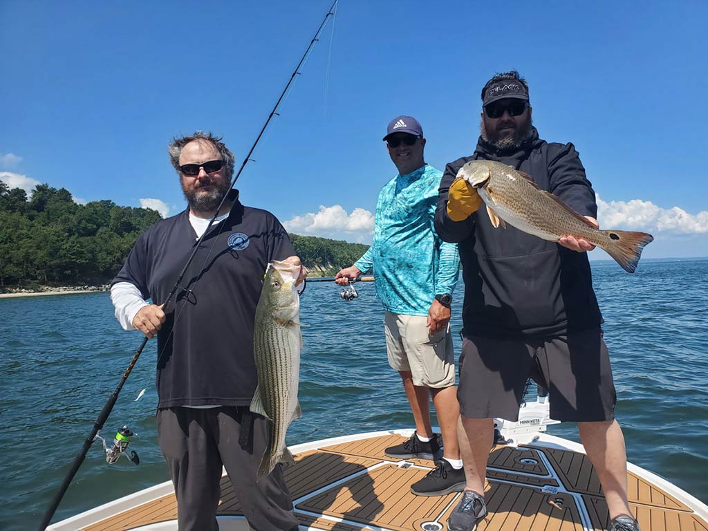 Трое мужчин на прибрежной рыболовной лодке в ясный день, демонстрируют улов полосатого басса и красного окуня, держа удочки.
