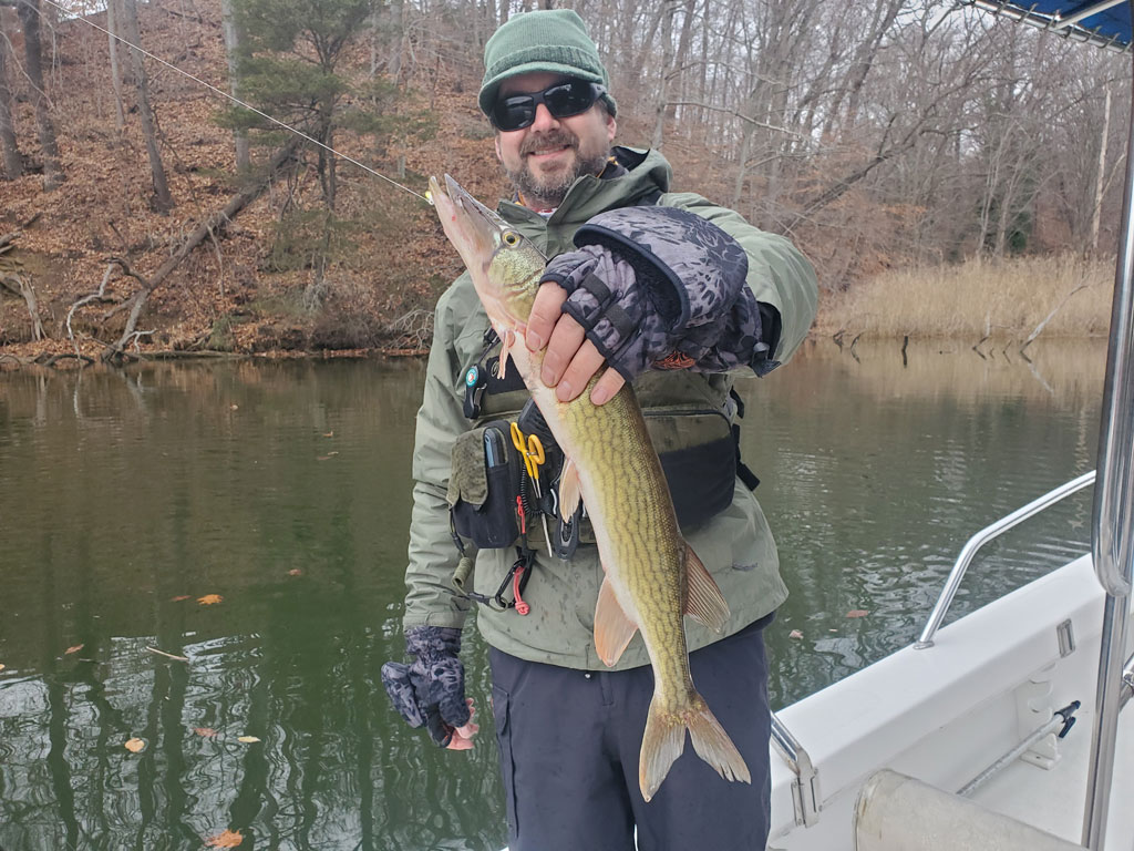 Мужчина в зимней рыболовной экипировке держит щуку на спокойной реке в холодный день.
