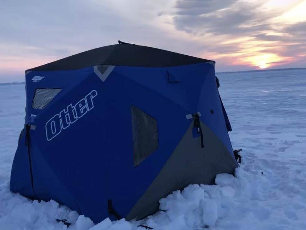 A blue Otter brand ice fishing shelter sitting on a snowy, frozen lake at sunset, with cloudy skies in the background.