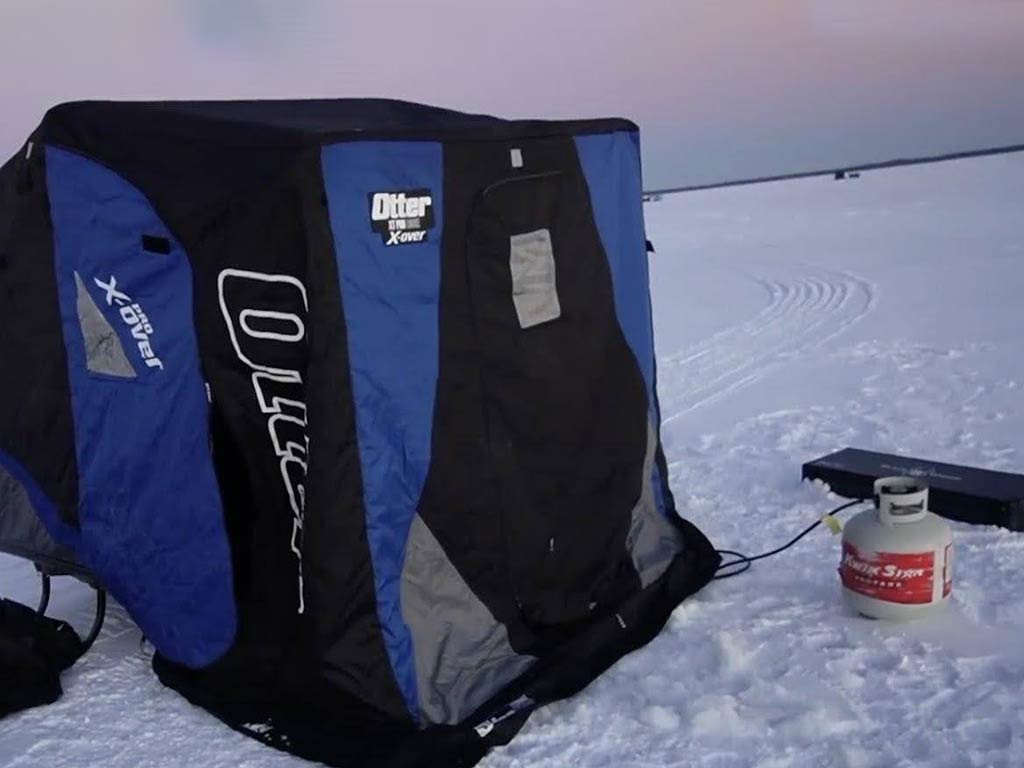 A blue and black portable ice fishing shelter sits on a snowy, frozen lake next to a propane tank and heater.